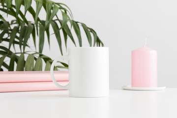 Mug mockup with books and a candle on a white table and a palm plant.