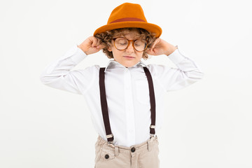 Beautiful boy with curly hair in white shirt, brown hat, glasses with black suspenders stands isolated on white background