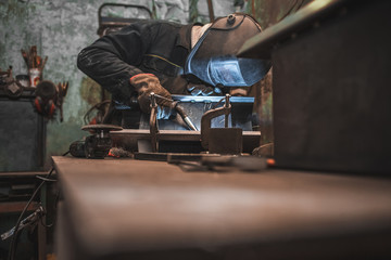 Worker in a welder mask works in a workshop for welding iron. Man makes iron products. Guy works with a welding machine using a mask to protect his eyes from dangerous rays