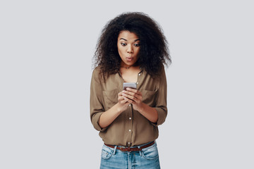 Surprised young African woman using smart phone and making a face while standing against grey...