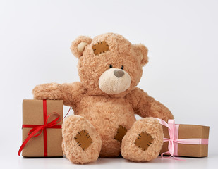 big beige teddy bear with patches, a stack of gifts in boxes wrapped in brown paper, tied with silk ribbon on a white background
