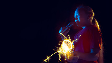 Caucasian girl's portrait on dark studio background in neon light. Beautiful female model with speaker and sparkler. Concept of human emotions, facial expression, sales, ad, hobby, dream, music. Flyer