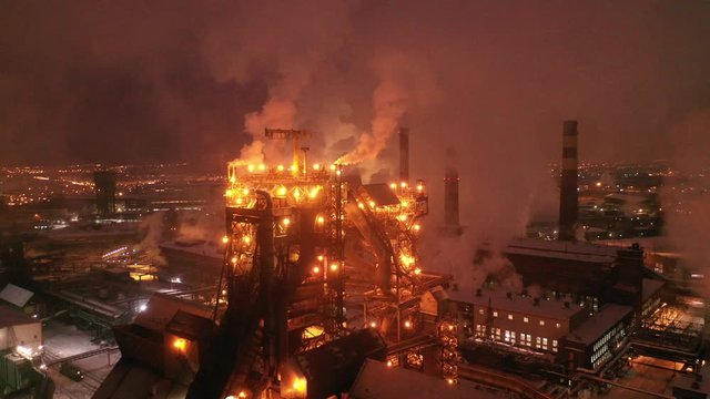 Aerial view of steel plant at night with smokestacks and fire blazing out of the pipe. Industrial panoramic landmark with blast furnance of metallurgical production. Concept of environmental pollution