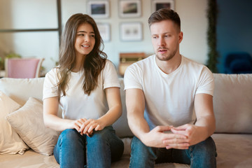 young couple man and woman sitting on the sofa both looking embarrassed , grimacing mood - obrazy, fototapety, plakaty