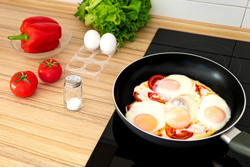 Traditional breakfast dish - fried eggs with tomato and pepper. Frying pan with hot fried eggs with tomatoes on black hob. Fresh lettuce, tomato, bell pepper and salt shaker on table.