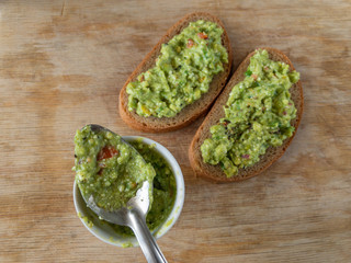 Top view on small cup and  spoon with guacamole next to sandwiches lying on a wooden table