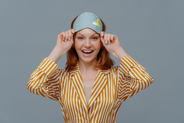 Ginger female model wears sleepmask and striped pajamas, awakes in good mood, realizes today is...