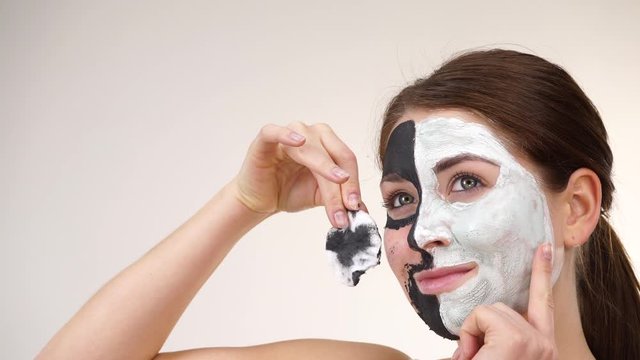 Woman with clay carbo black mask on one half face and white mud on second half. Girl removing cosmetic scrab with cotton swab. Female taking care of oily complexion. Skincare
