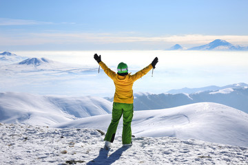Happy woman in yellow ski jacket standing with hands up on the pick of mountain Tegenis. View of...