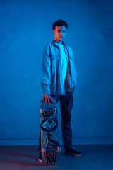 Caucasian young skateboarder in casual posing on dark neon lighted blue background. Confident and playful male model. Concept of hobby, healthy lifestyle, youth, action, movement, modern art style.