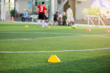 Obraz premium Marker Cone on green artificial turf with blurry kid soccer players are training background. Soccer academy.