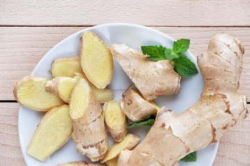 Fresh ginger root sliced and mint leaves on a white plate and a light wooden background. Close-up, top view.