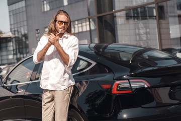 Fototapeta premium Mode of Transport. Man with long hair in sunglasses standing near electric car lighting cigarette looking aside cool