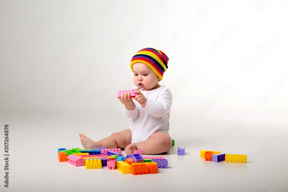 Wall mural baby boy in white bodysuit and multicolored hat playing with a multicolored plastic constructor on a white background isolate, child development concept, space for text