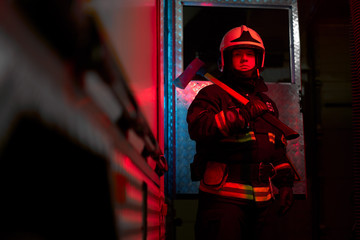 Obraz na płótnie Canvas Close up firefighter man with hammer on his shoulder standing near fire engine