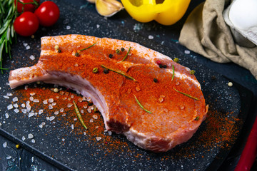 Raw pork steak on the bone sprinkled with red pepper near rosemary, corn, eggs and bell pepper.