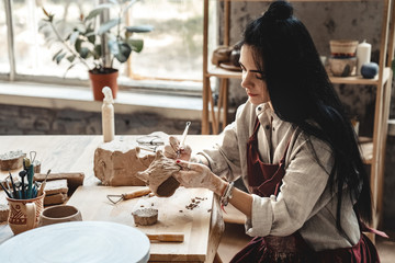 Craftsperson Concept. Young woman making pottery indoors making shape for clay using tool modeling...