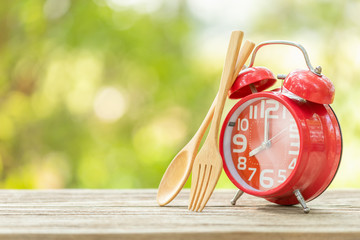 Red alarm clock, Fork, and spoon on wooden table with green outdoor nature blur background. Eight...