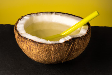 half of coconut with a plastic cocktail straw lies on a stone tray on a yellow background