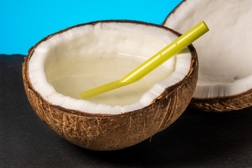 two half of coconut with a plastic cocktail straw lies on a stone tray on a blue background