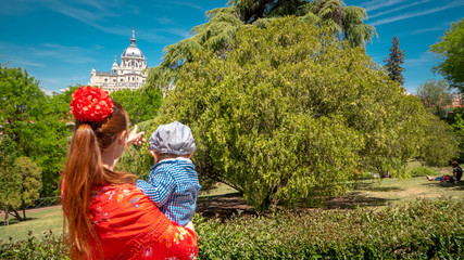 A young caucasian woman chulapa is holding her baby chulapo in traditional dresses with Almudena cathedral during San Isidro, the spring festival in May in the downtown of Madrid, the capital of Spain
