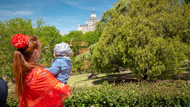 A young caucasian woman chulapa is holding her baby chulapo in traditional dresses with Almudena cathedral during San Isidro, the spring festival in May in the downtown of Madrid, the capital of Spain