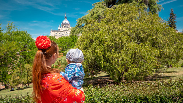 A young caucasian woman chulapa is holding her baby chulapo in traditional dresses with Almudena cathedral during San Isidro, the spring festival in May in the downtown of Madrid, the capital of Spain