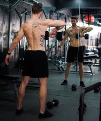 Fototapeta premium Caucasian Shirtless Tattooed Man Exercising Traps Using Dumbbells