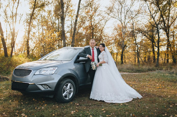 Happy newlyweds are hugging on the background of the forest with yellow trees, standing near the car. Wedding autumn portrait of a stylish bride and groom on the nature. Photography, concept.