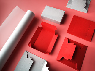 Creative crafts, pink background with roll of tracing paper, flat templates for flat cardboard gift boxes and ready box. Paper craft objects in pink, red and grey.
