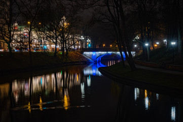 Fototapeta na wymiar Riga's City Canal by night with the city lights reflecting in the water in foggy evening and small bridge lightened with blue light during Staro Riga festival