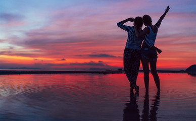 Two girlfriends hugging up and enjoying a rose/pink sunset sky on the sea beach on the Samui...