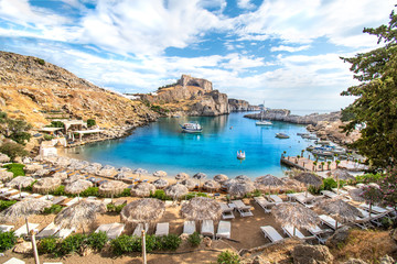 Lindos – view of St. Paul bay, beach with sun beds and umbrellas, acropolis of Lindos in background (Rhodes, Greece)