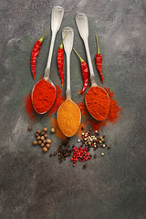 Various types of pepper in spoons on a dark grunge background. Paprika, chili, smoked paprika, hot pepper, peppercorns. Top view, flat lay, copy space.