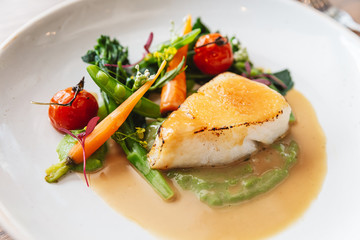 Close-up Pan seared Cod fish fillet with tomato, bean and carrot in gravy sauce. Served in a white plate with a silver knife and fork.