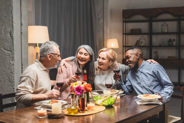 smiling multicultural friends hugging and holding wine glasses during dinner