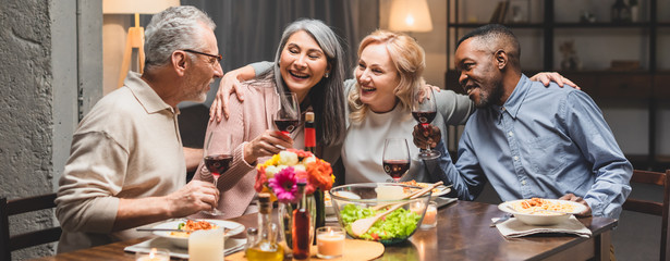 panoramic shot of smiling multicultural friends hugging and holding wine glasses during dinner