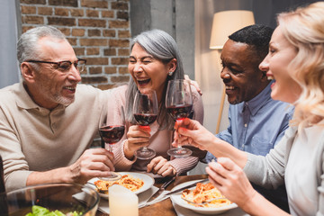 smiling multicultural friends talking and clinking with wine glasses during dinner
