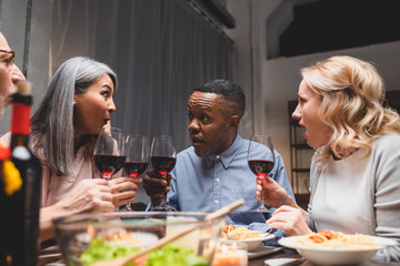 shocked multicultural friends talking and holding wine glasses during dinner