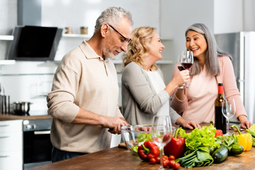 smiling multicultural woman clinking and man cutting lettuce in kitchen