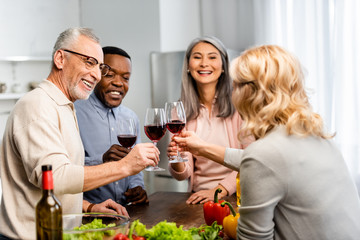 smiling multicultural friends clinking with wine glasses in kitchen