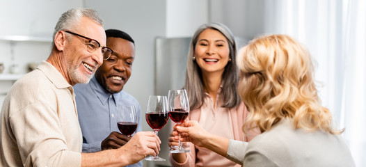 panoramic shot of smiling multicultural friends clinking with wine glasses in kitchen