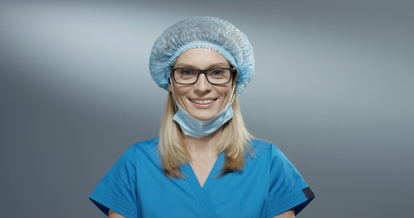 Portrait shot of the young attractive blond Caucasian female physician in the blue hat and glasses taking off her mask from the mouth and smiling happily. Close up.