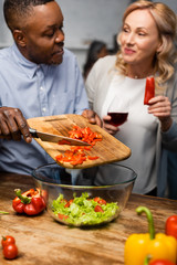 selective focus of smiling african american man adding cutting bell pepper to bowl and woman holding wine glass and bell pepper
