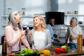 selective focus of smiling multicultural friends holding wine glasses in kitchen