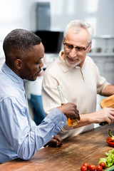african american man holding beer and his smiling friend cooking in kitchen