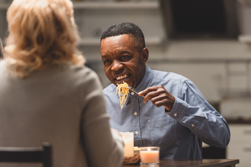 selective focus of smiling african american man eating pasta and talking with friend during dinner