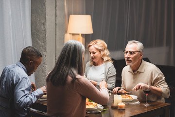 selective focus of smiling man and woman talking with multicultural friends and eating during dinner