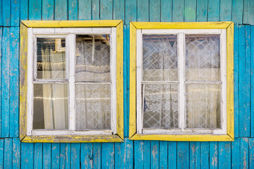 Fragment of wall of old wooden house with two windows with cracked paint on frames and holes between boards. Cornice with curtain behind glass. Rustic style