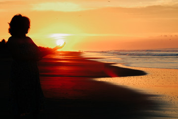 silhouette of a unrecognizable woman on the beach at sunrise. She's pretending to hold the sun with her hand.